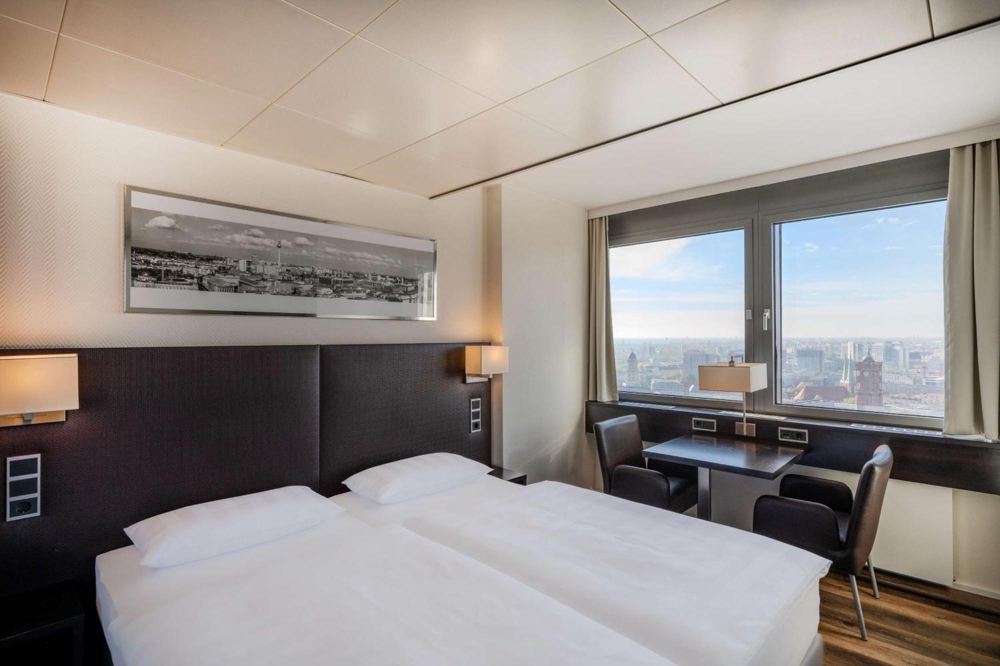 Park Inn By Radisson Berlin Alexanderplatz Buitenkant foto The photo shows a modern hotel room featuring two single beds with white linens and dark headboards. There is a large window that provides natural light and a view of the cityscape outside. The room is equipped with a small table and two black chairs